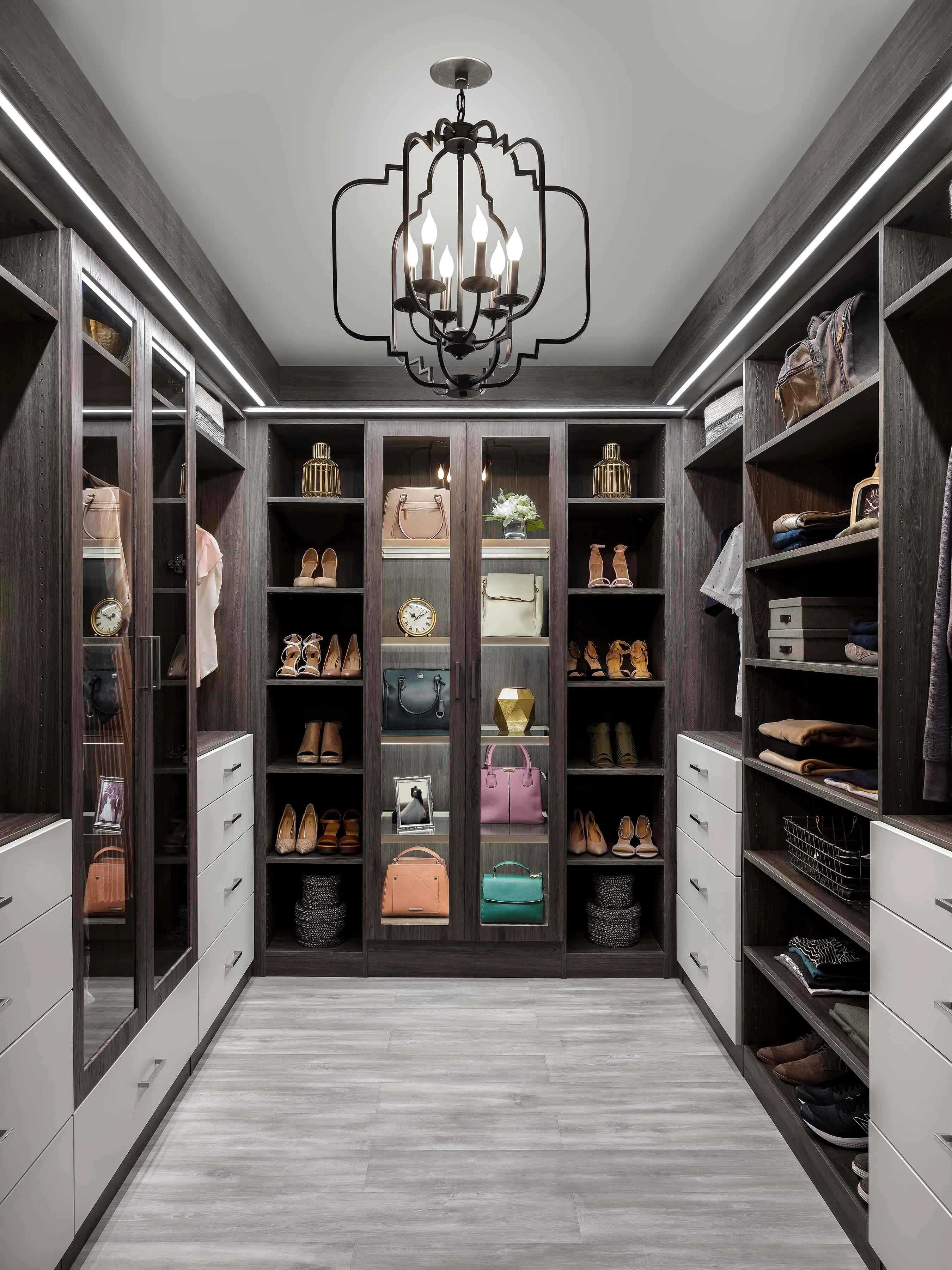 Luxury dark wood walk-in closet with illuminated displays, white drawers, shoes, handbags, and a geometric chandelier.