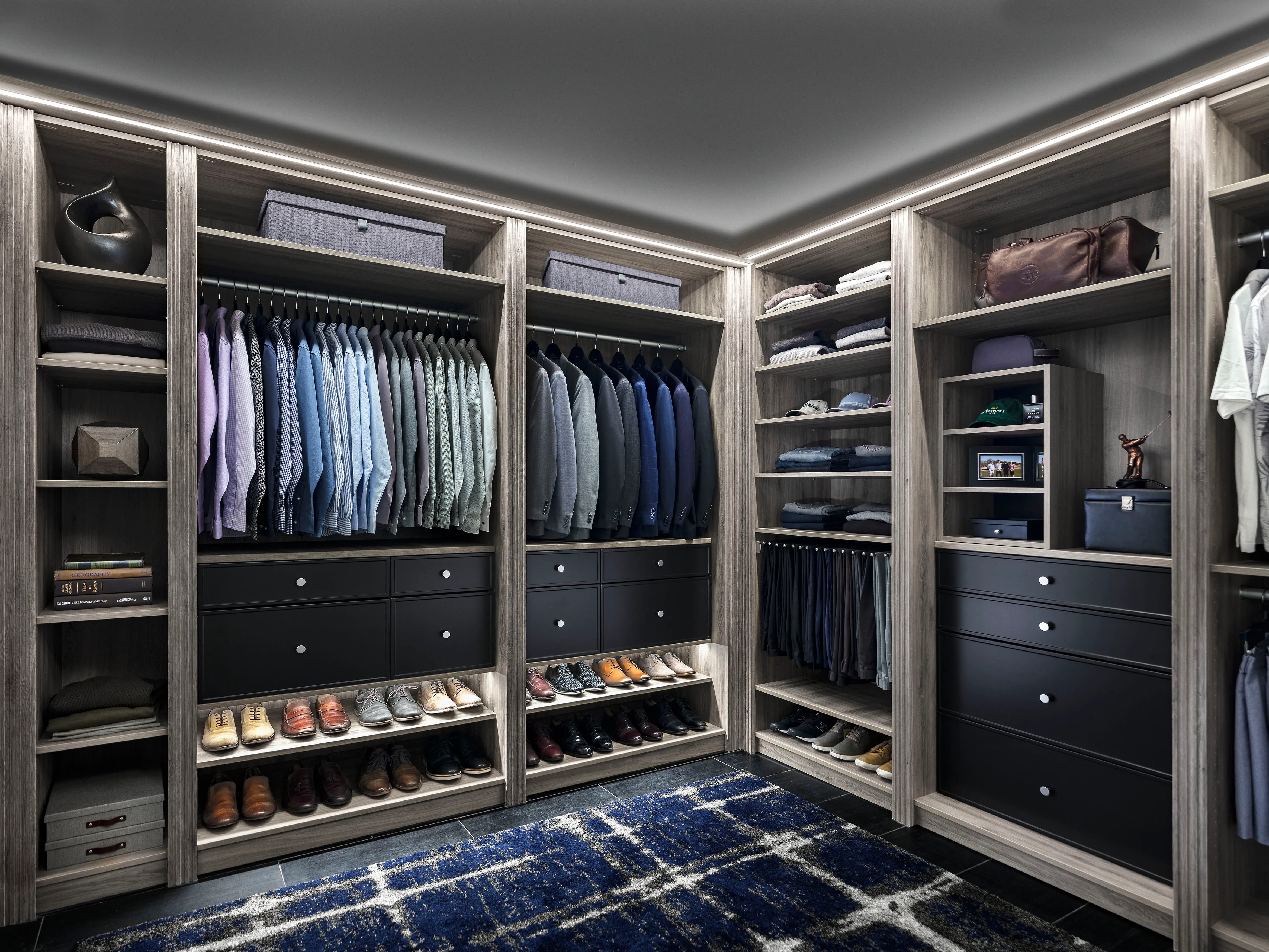 Men's gray wood walk-in closet with black drawers, hanging clothes, shoe shelves, and integrated LED lighting.