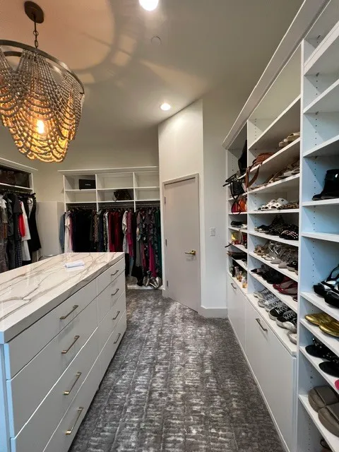 White walk-in closet with a marble-look drawer island, shoe shelves, hanging clothes, and a beaded pendant.