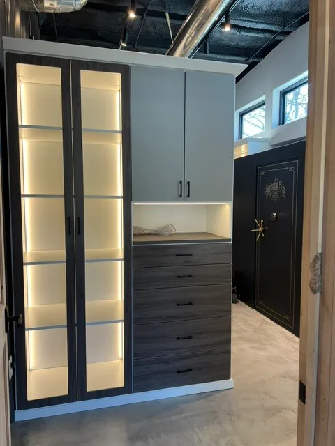 Dark display cabinetry with lit glass doors, gray drawers, upper cabinets, and a safe in the background.