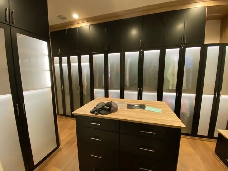 Black walk-in closet with illuminated translucent glass doors and a central drawer island with a wood top.