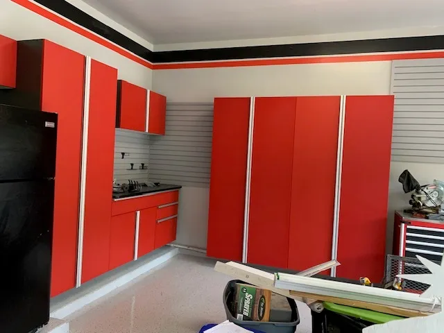 Red garage cabinets with silver trim, black worktops, gray slatwall, and project materials in the foreground.