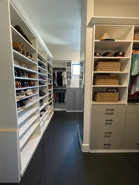 Narrow white walk-in closet with long shoe shelves, woven baskets, hanging clothes, drawers, and dark flooring.