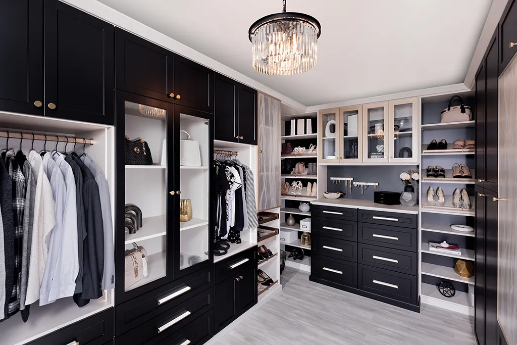 Black and white luxury walk-in closet with glass cabinets, open shoe shelves, drawers, brass hardware, and a crystal chandelier.