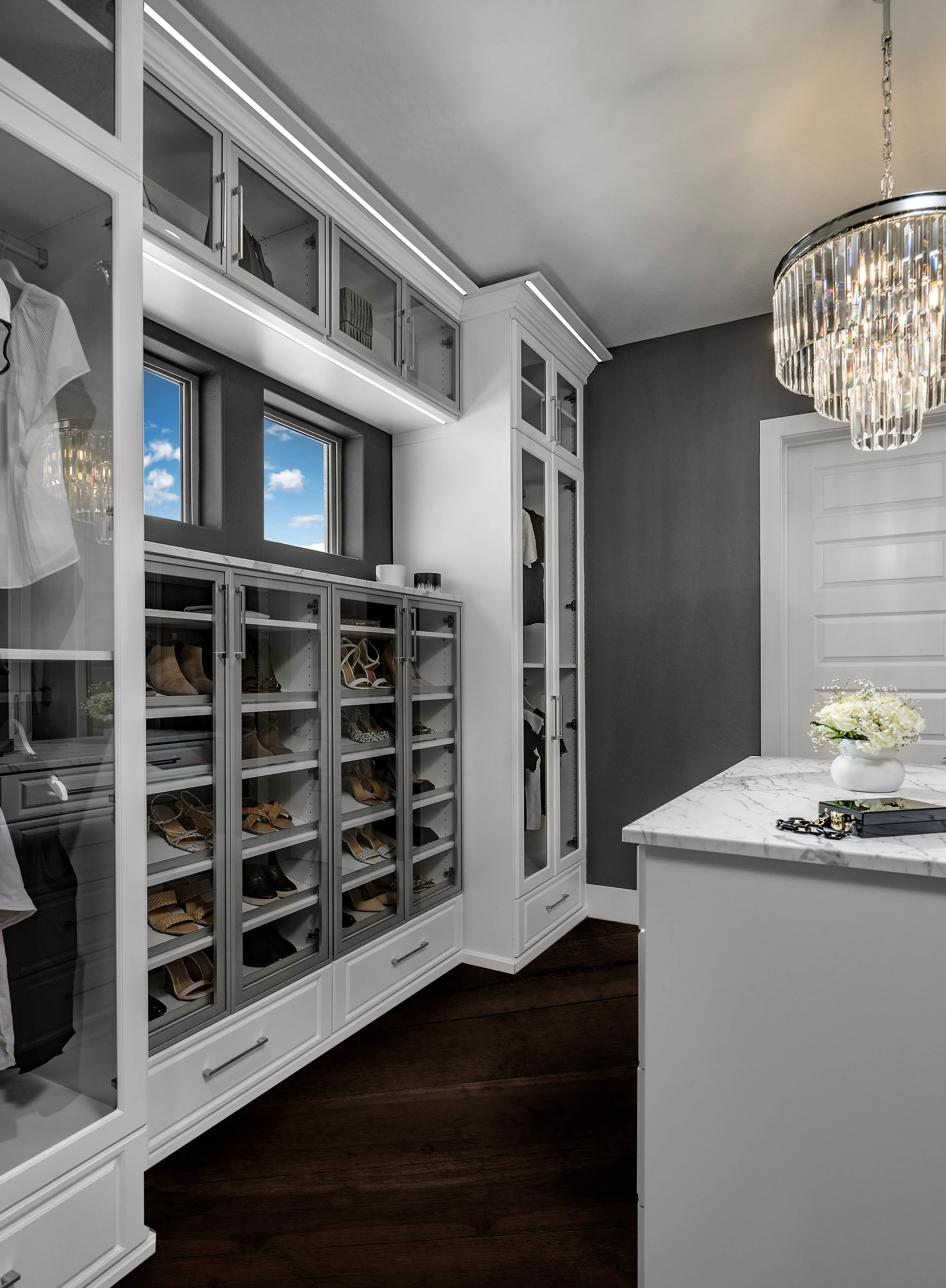 Luxury white closet with metal-framed glass cabinets, shoe displays, marble island, dark walls, and a crystal chandelier.
