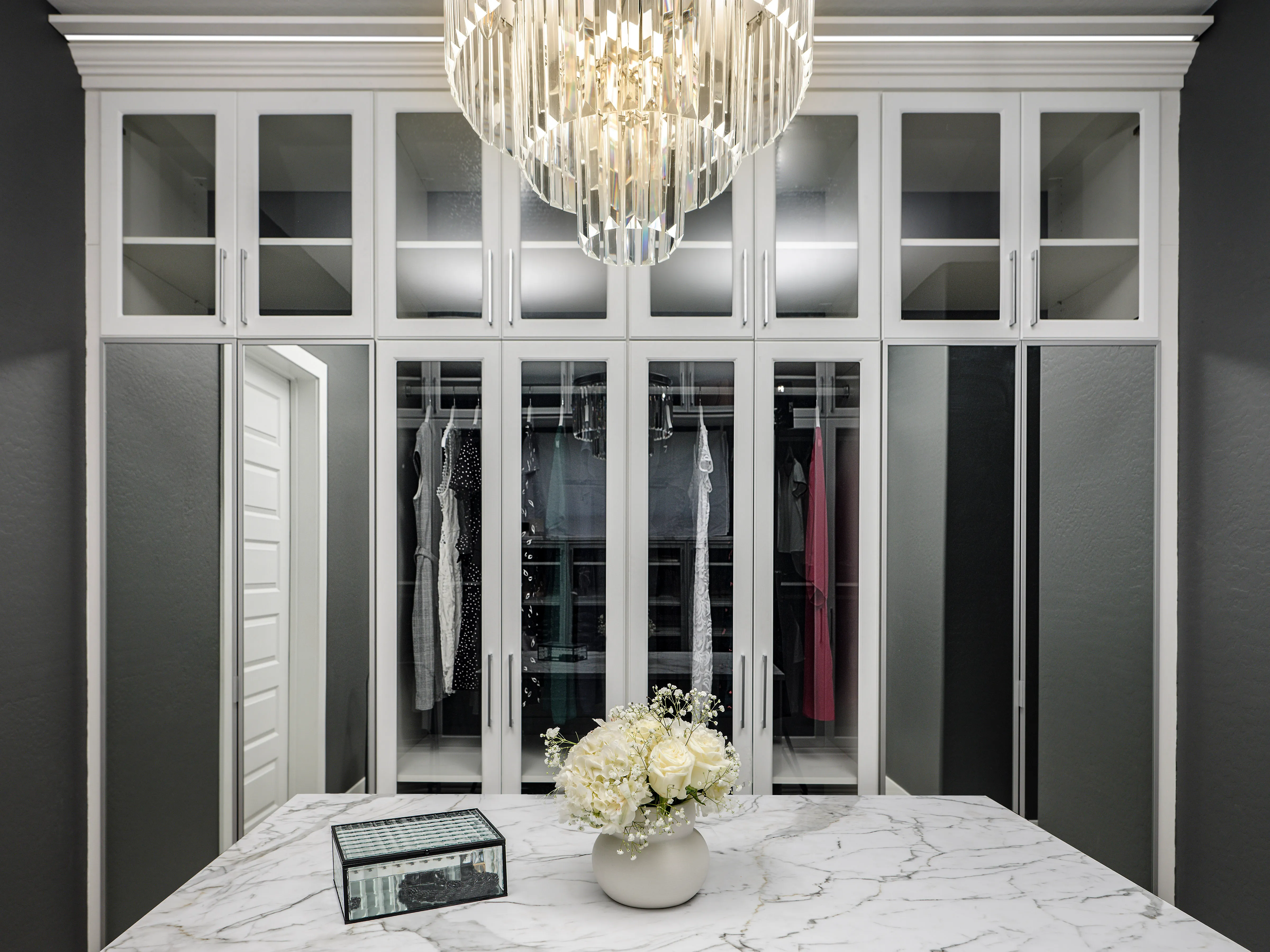 Symmetrical white closet with upper glass cabinets, tall glass and mirrored doors, a marble island, and crystal chandelier.